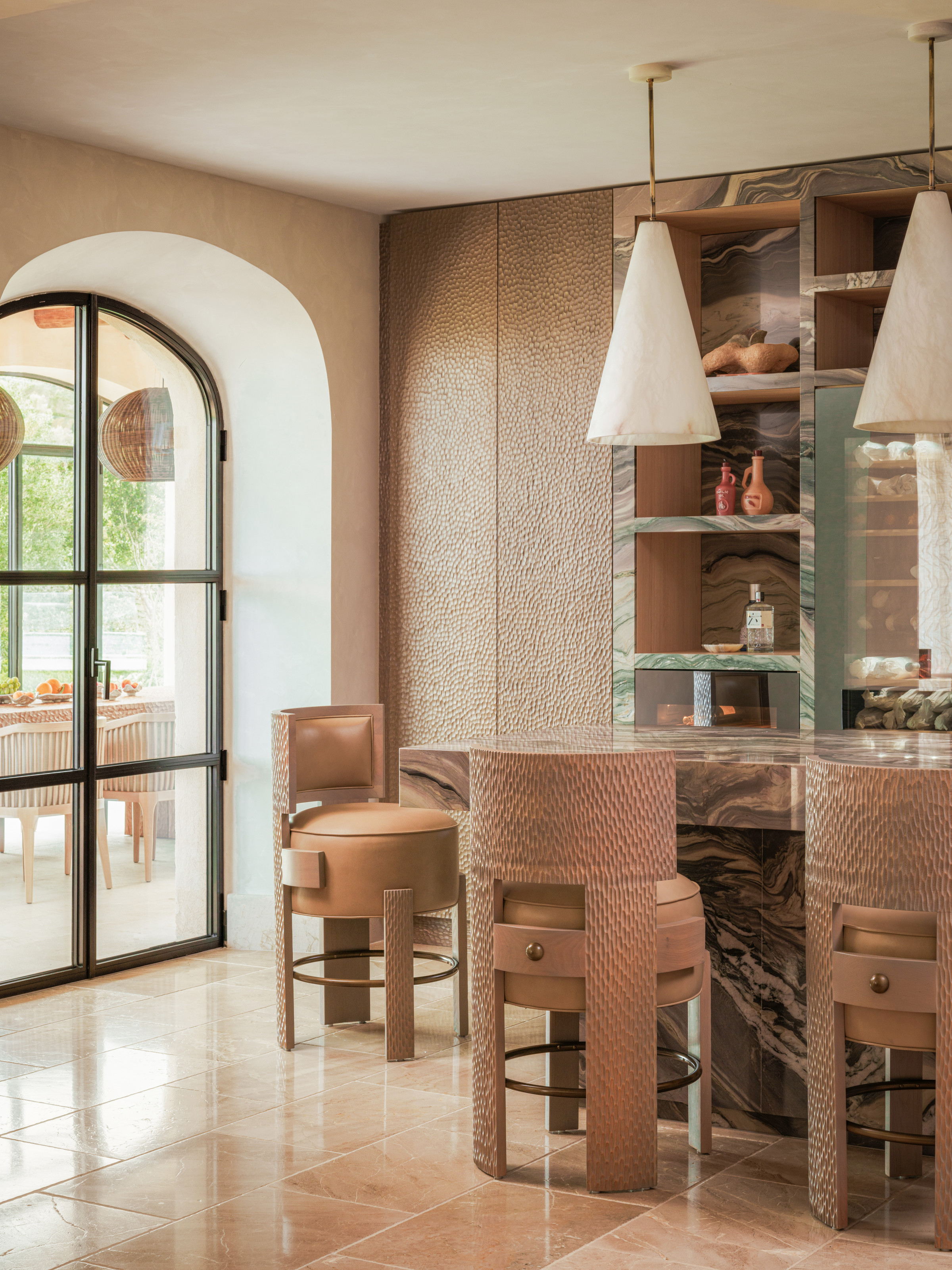 Kitchen with brown textured cupboard and green marble shelving, brown marble island, brown textured barstools and oversized white marble pendants
