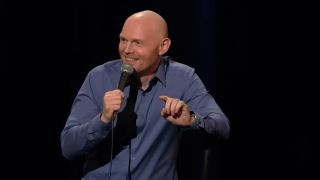 Bill Burr in a blue shirt doing standup, pointing and smiling
