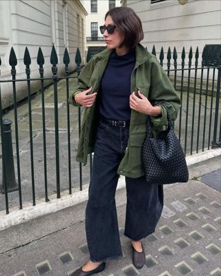 Fashion influencer Lucy Alston in London wearing a chic fall outfit with barrel leg black jeans, mesh flat shoes, a green jacket, and Rylan woven leather bag.