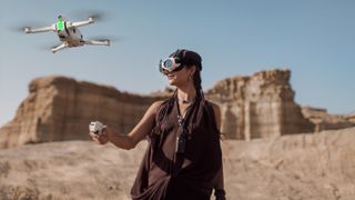 A woman wearing a brown dress and goggles holds a remote and looks up at a white drone with green lights flying in a vast, dry canyon.