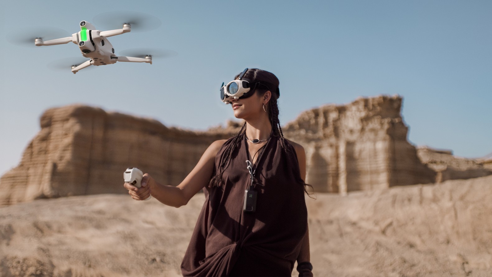 A woman wearing a brown dress and goggles holds a remote and looks up at a white drone with green lights flying in a vast, dry canyon.