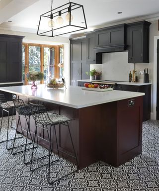 A kitchen with black cabinets, a white kitchen cabinet with burgundy back panels and three metal chairs in front of it, and black and white mosaic patterned tiles