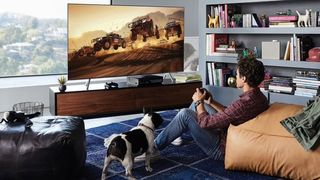 En mand sidder på stuegulvet sammen med en hund og spiller på et Samsung-tv foran et stort vindue