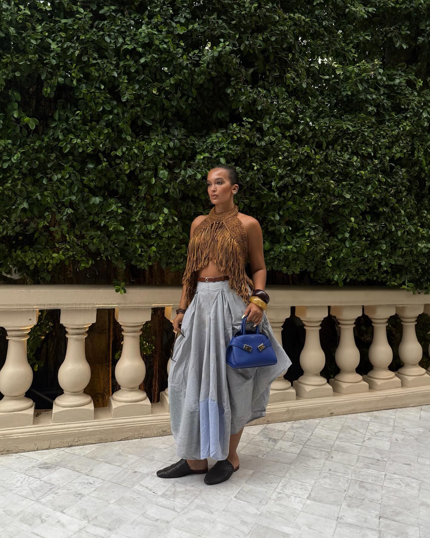 Oumayma Elboumeshouli holding a sapphire blue top-handle bag against pleated gray maxi skirt.