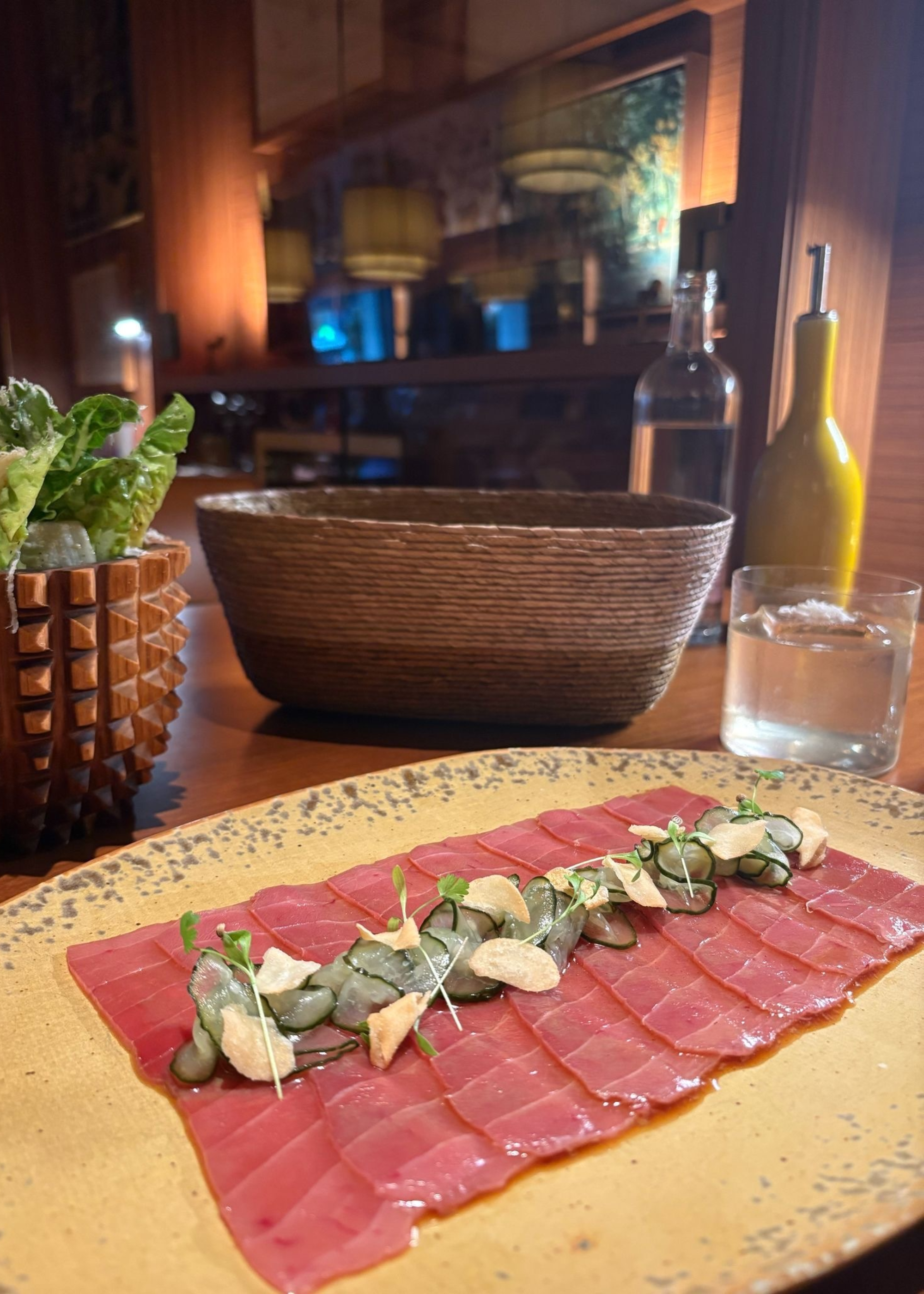 A plate of red tuna with seaweed and algae beside a bowl of Caesar salad, a bread basket, a cocktail and a bottle of olive oil