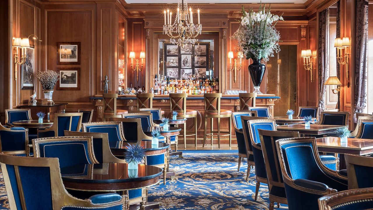 Image of a formal and traditional style dining room. The walls are wooden and there are blue and gold carpets. The tables and chairs are blue and gold as well.