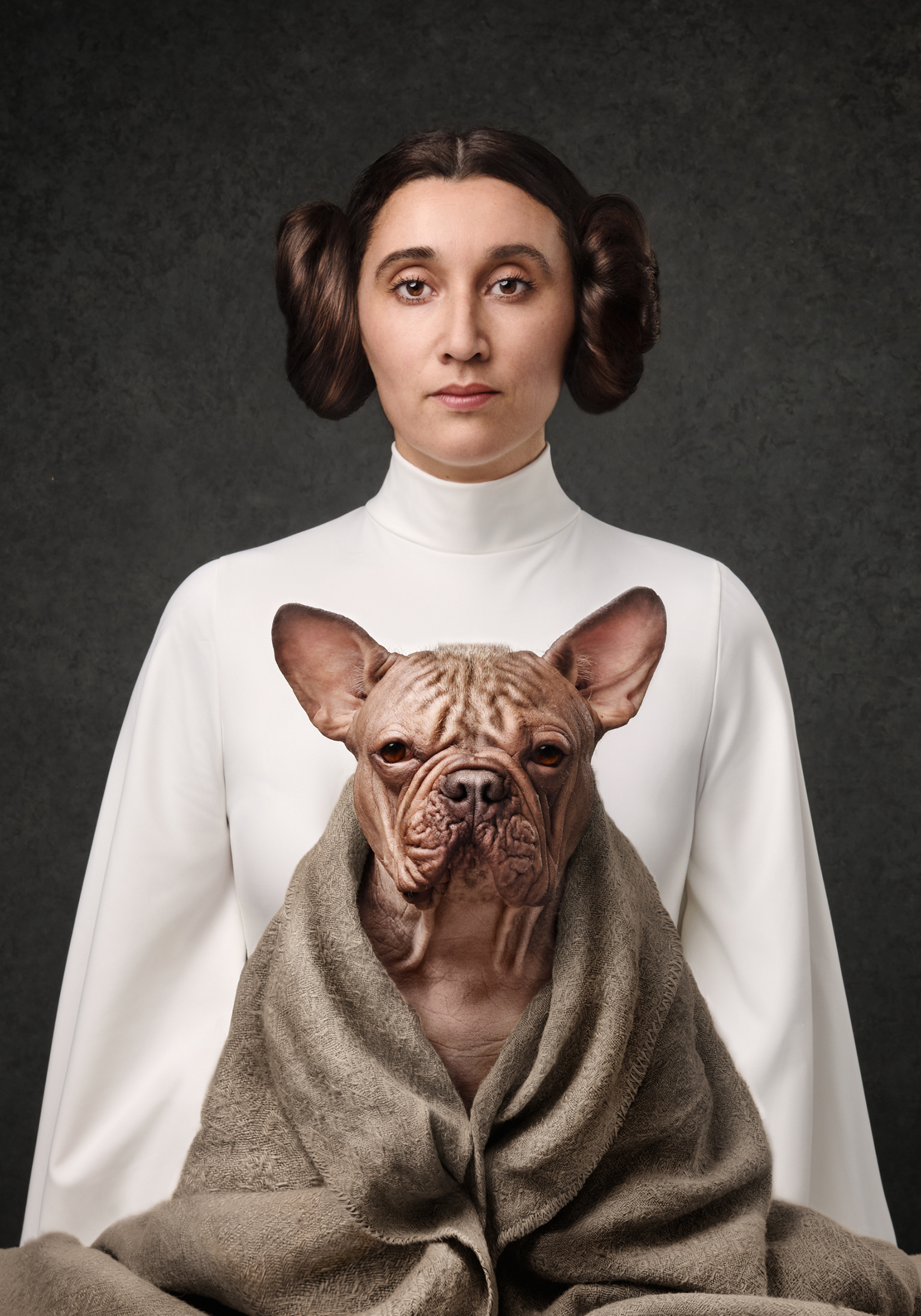 A person in a white turtleneck sweater holds a French Bulldog wrapped in a brown blanket, against a dark background