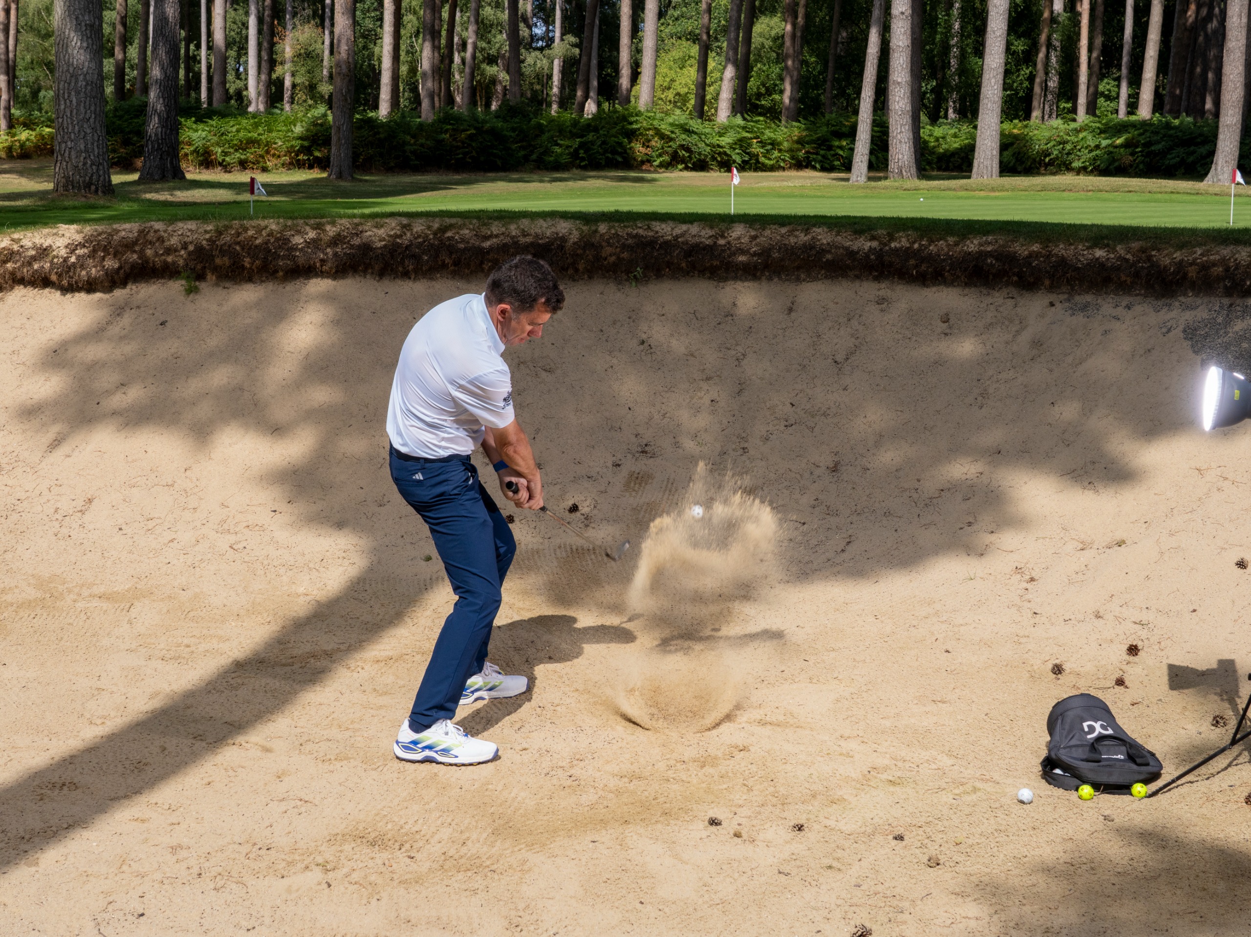 Dan Grieve hitting a bunker shot with the club just past impact and the ball appearing out of a splash of sand