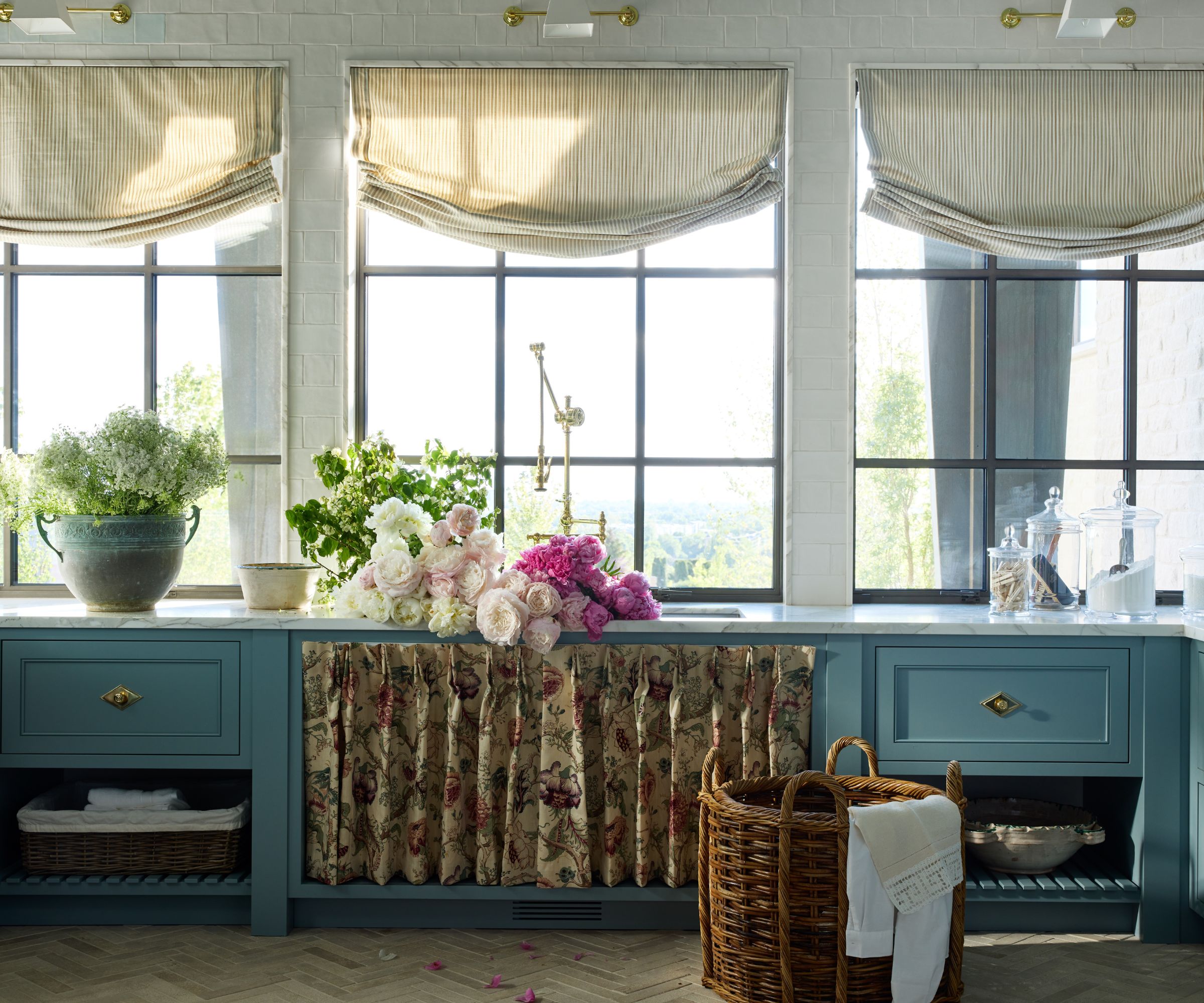 Laundry room with powder blue cabinetry, a floral skirted cabinet, a bouquet on the countertop beside a sink, three large windows with roman striped blinds, and a rattan laundry hamper.