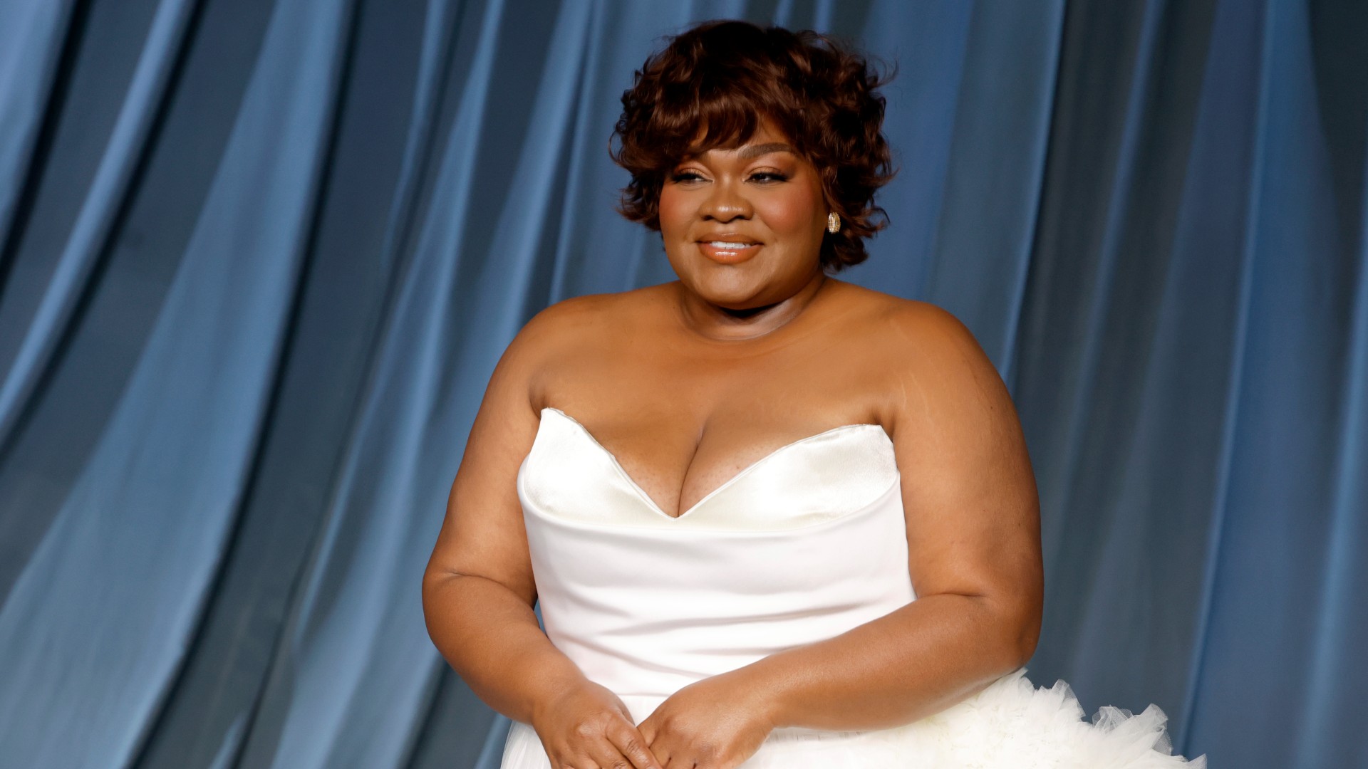 a black woman with short brown hair poses in a white strapless gown in front of a blue backdrop