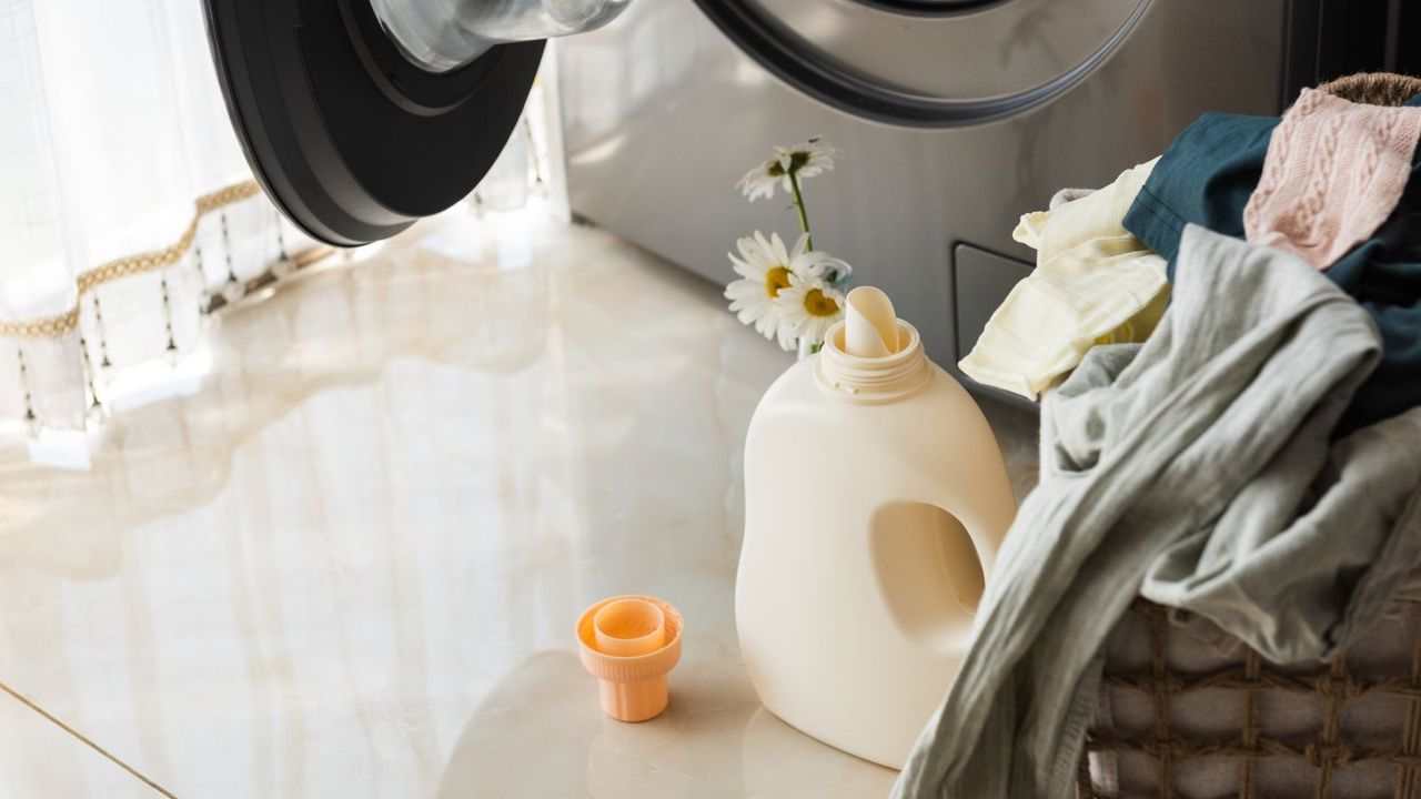 A labelless bottle of laundry detergent with its lid off and some flowers stuck in the top nect to awashing machine and a basket of laundry