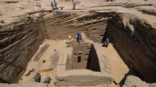 This photo shows a bird's eye view of a tomb as it is being excavated in Egypt. There are 5 people scattered inside the big hole, which requires a long ladder to get to. Above there are 5 people also doing excavation work.
