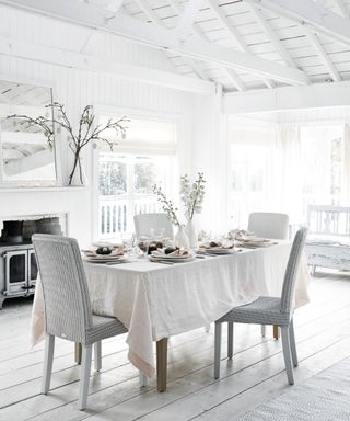 All-white dining room space, white paneling walls and wooden beams, rectangular dining table with off-white table cloth, four upholstered gray dining chairs, white fireplace with stove, mirror placed above, light and bright space