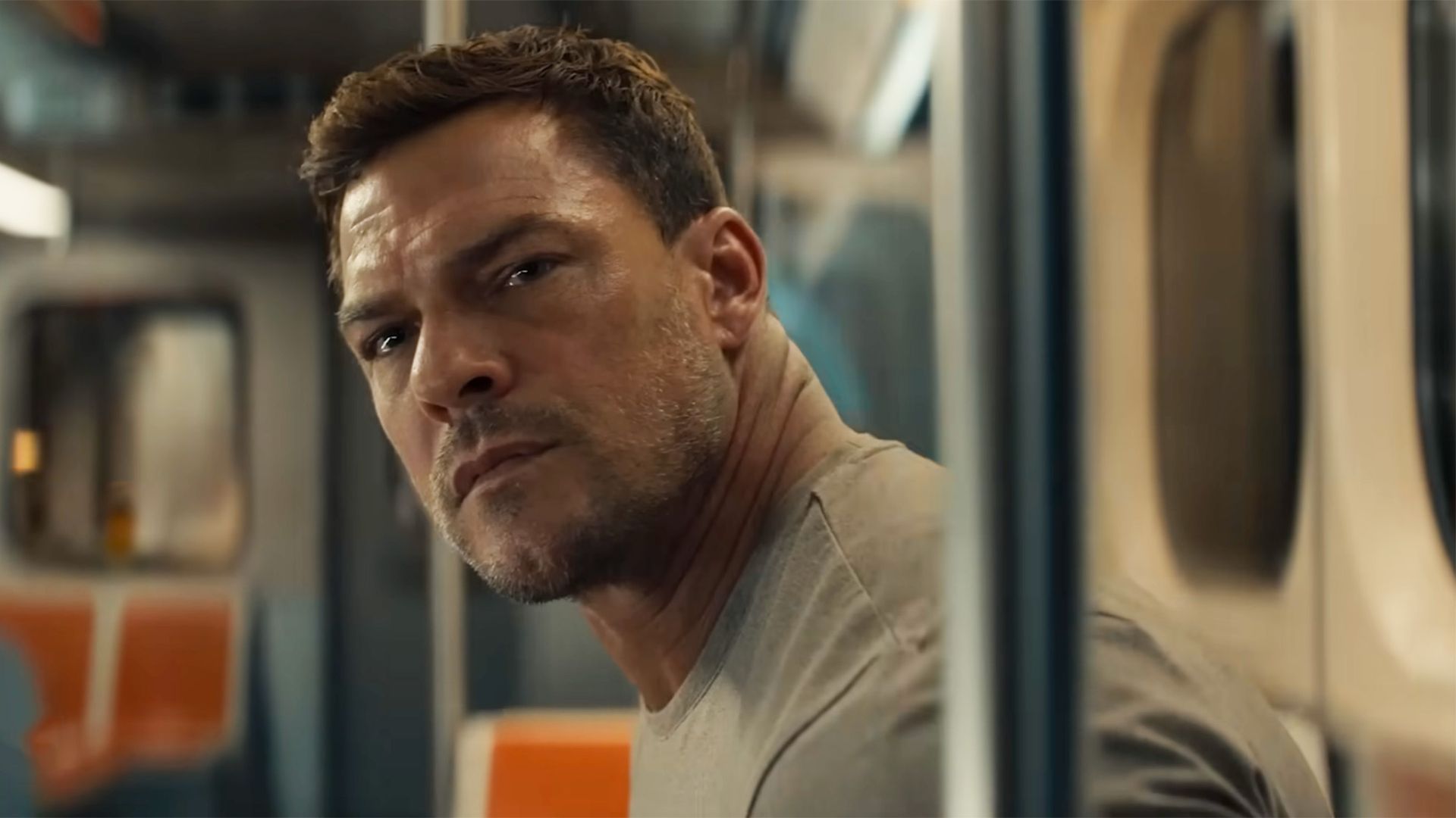 Alan Ritchson looks concerned in a tan tee on public transportation while staring at the camera in Reacher.