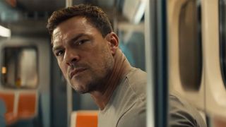 Alan Ritchson looks concerned in a tan tee on public transportation while staring at the camera in Reacher.