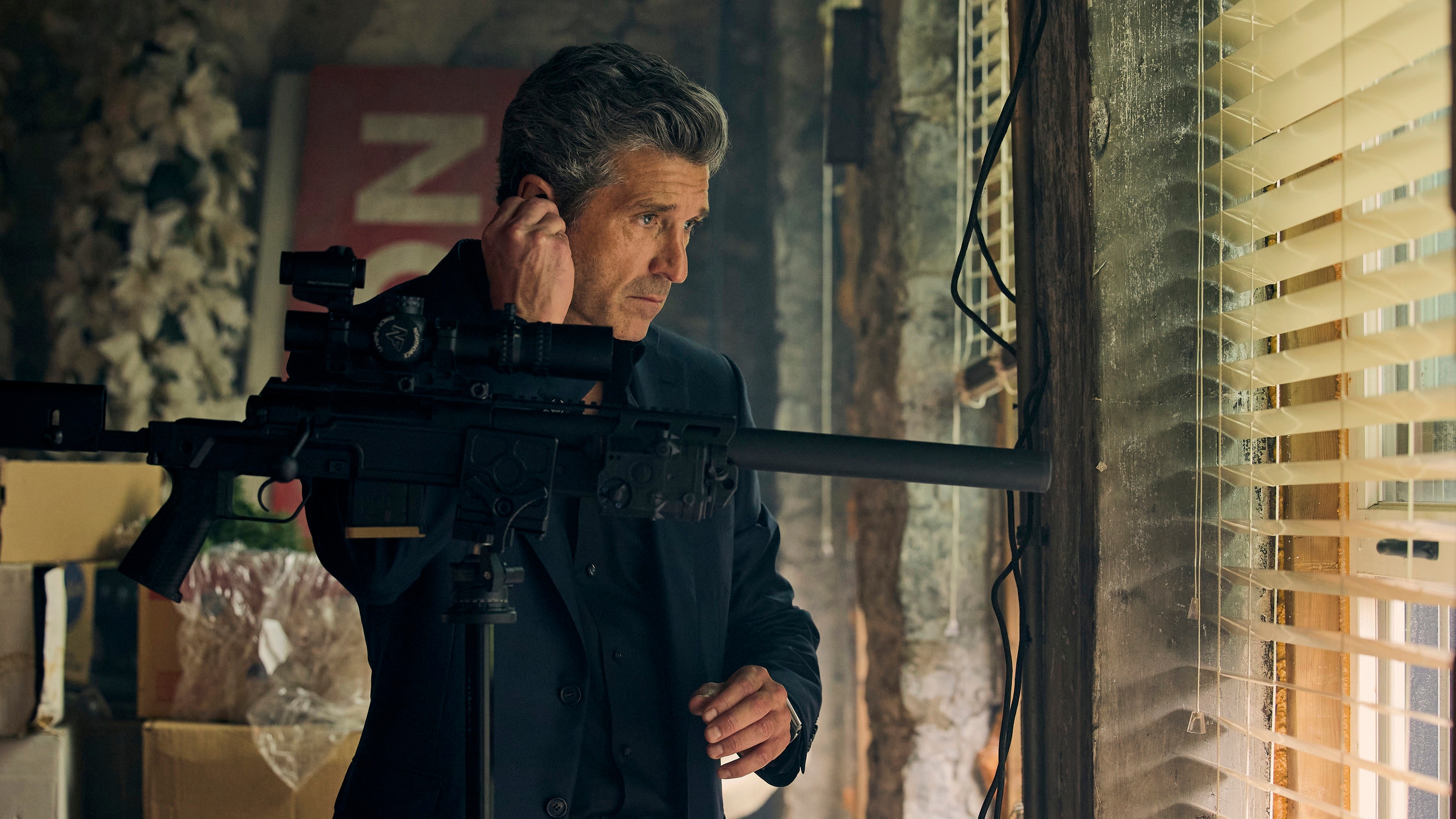 Patrick Dempsey as Angelo looking out of a window while adjusting something in his ear.
