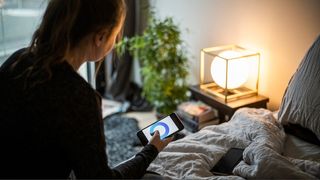 Woman sitting on AI mattress with smart phone in hand checking her sleep report. Grey bedsheets, round bedside lamp and a plant in background.