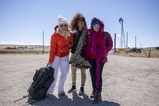 Trailblazers: A Rocky Mountain Road Trip stars Emily Atack, Melanie Brown and Ruby Wax stand on a dirt highway in a remote location in the USA, all wearing warm coats. Emily is dragging a backpack along the ground.