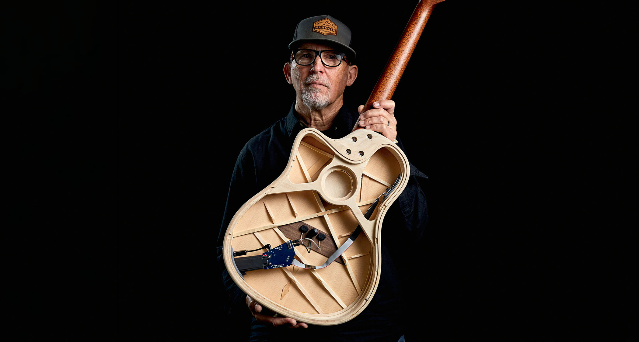 Lloyd Baggs wears a ballcap and spectacles and is pictured in a dark room with the groundbreaking AEG-1 acoustic-electric guitar.