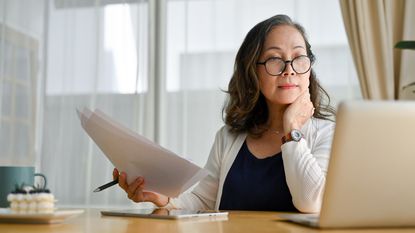 A woman holds paperwork and looks at her laptop while sitting at her desk.