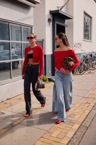 Two woman wearing pops of red. One woman is wearing a red cardigan that is opened with a black bandeau, black high-waisted jeans, red sandals, and a black bag. The other woman is wearing an off-the-shoulder red top, cheetah-print clutch, wide-leg jeans, and red ballet flats.