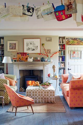 A classic country living room with orange chairs, pattered cream ottoman and fireplace