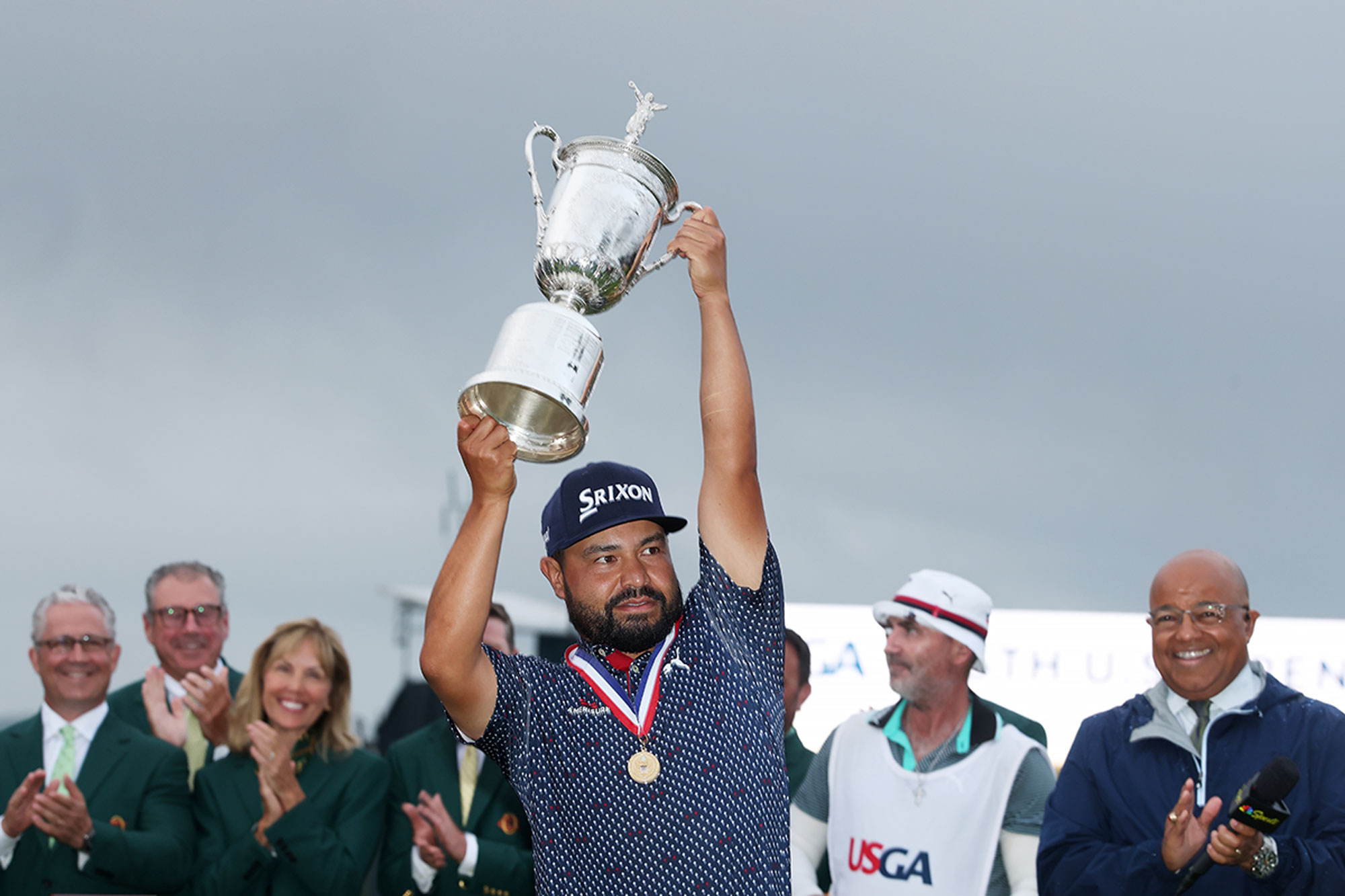 JJ Spaun raising the US Open trophy above his head, in front of his caddie and other onlookers, after winning the Major at Oakmont Country Club