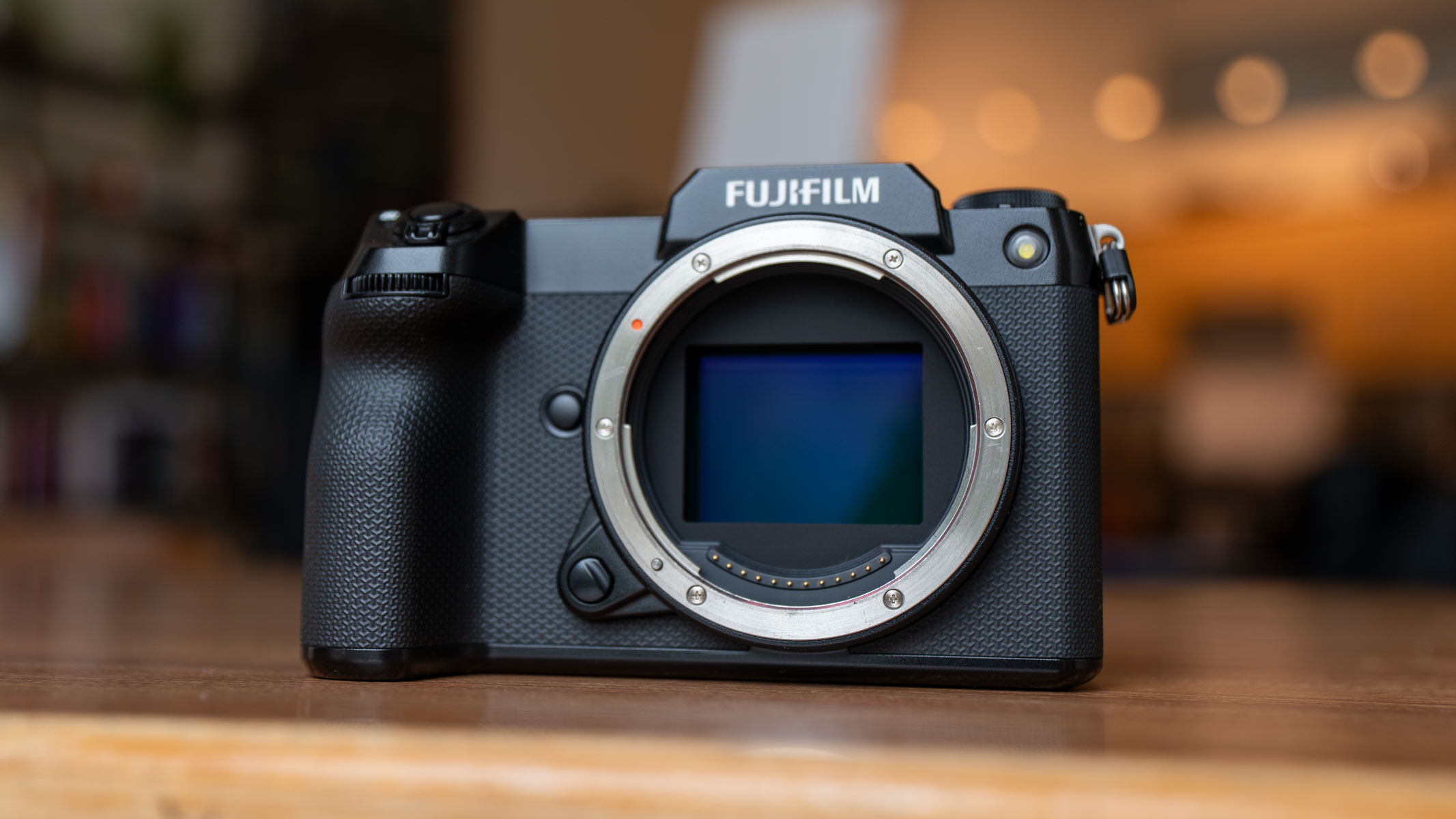A head-on shot of the Fujifilm GFX100S II showing its sensor in blue on a wooden table, with lights in the background.
