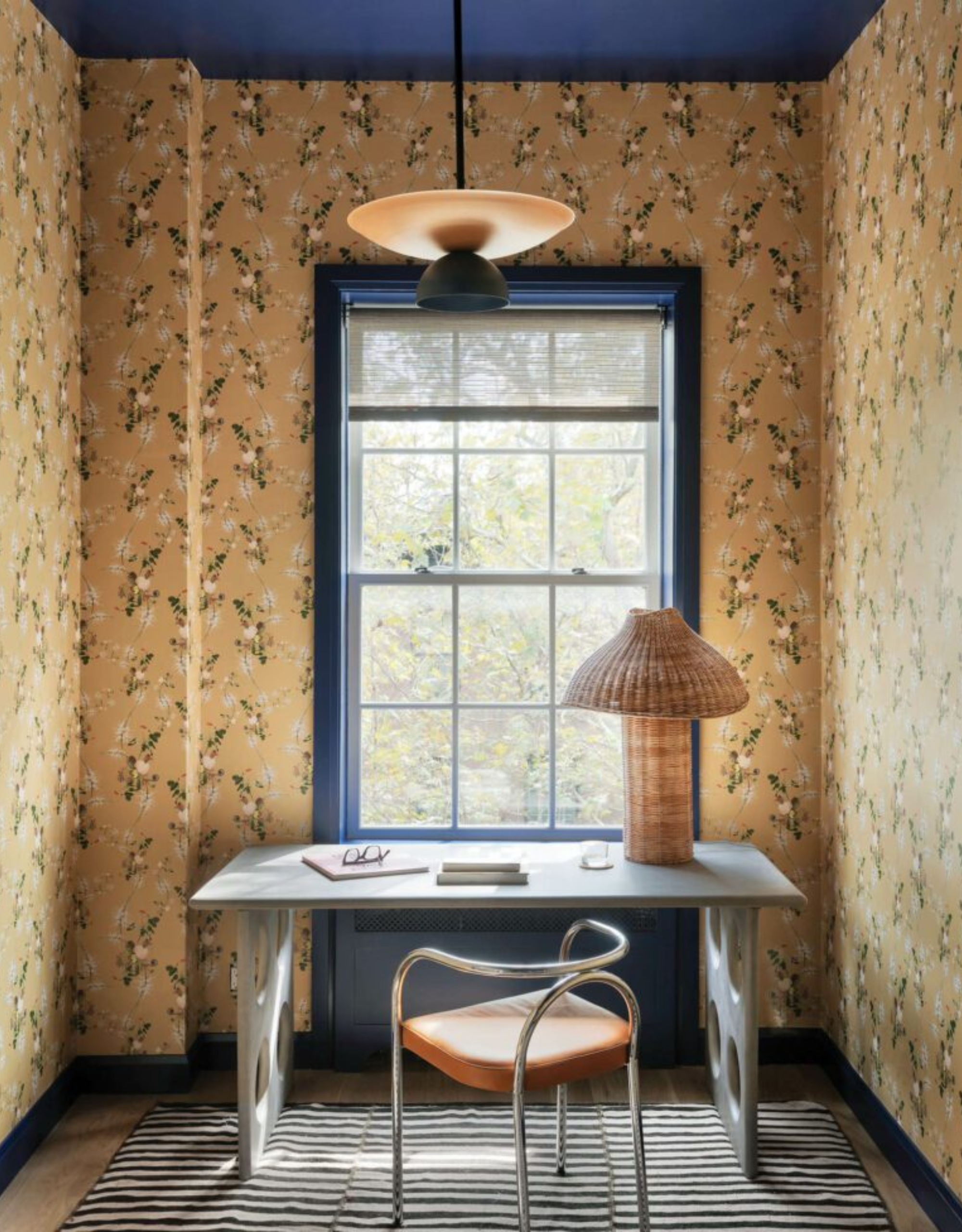 A small home office with patterned yellow wallpaper, blue trim around the window and a blue capped ceiling, a structured desk, a metal chair, and a woven lamp on a striped rug