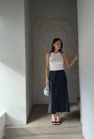 Hanukkah outfit idea is shown in a photo of a woman standing on concrete stairs wearing silver earrings, a white tank top, a black slip skirt, black mules, and a silver clutch bag