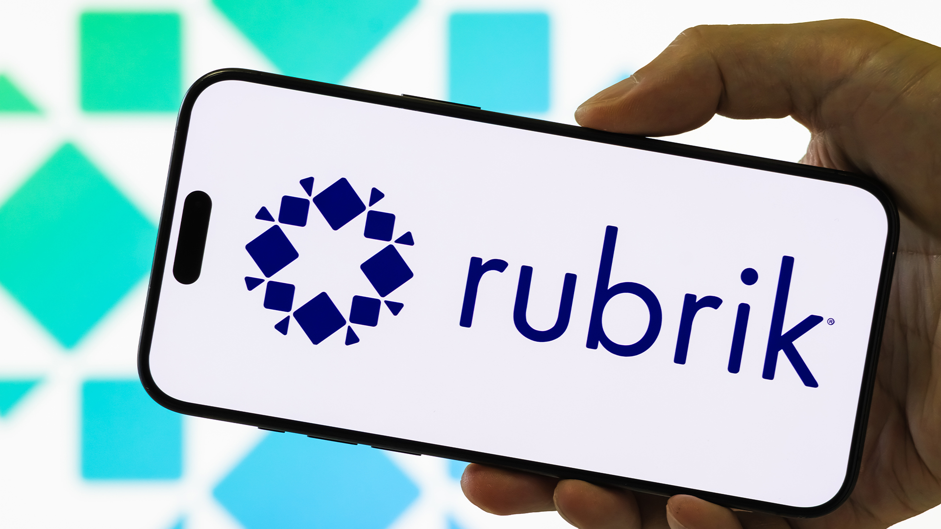 Rubrik logo and branding pictured on a smartphone screen held in a person's hand, with company logo in background. 
