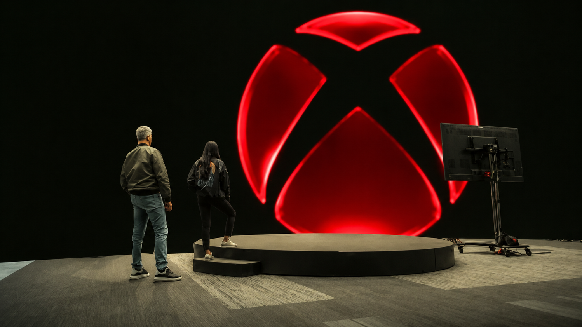 Xbox executives Asha Sharma and Matt Booty stand before a large illuminated Xbox logo in red, highlighting the company's recent struggles