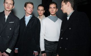 Five male models wearing looks from Canali's collection. They are wearing dark coats, white and grey jumpers and grey striped trousers