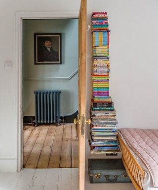 Floating bookshelf with colorful stack of assorted books between door and foot end of bed.