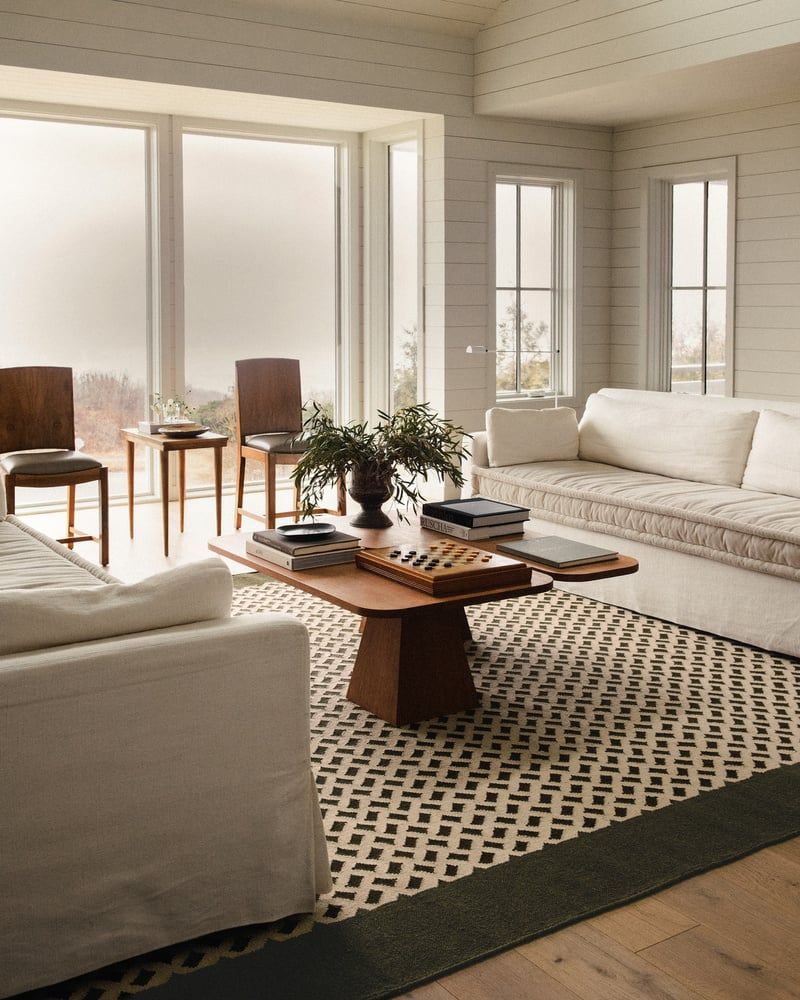 Image of a white living room with lots of windows and wood panelled walls. There are two white sofas facing each other with a wooden coffee table between them and a brown and white rug on the floor that has a basket weave pattern.