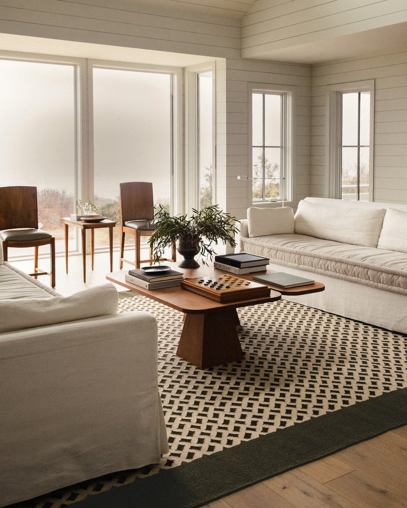 Image of a white living room with lots of windows and wood panelled walls. There are two white sofas facing each other with a wooden coffee table between them and a brown and white rug on the floor that has a basket weave pattern.
