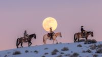Three cowboys on horses wearing ten-gallon hats cross from left to right across a snowy hill with a large yellow full moon in a pink night sky behind them