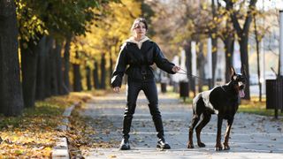 Young Woman Wearing Black Leather Clothes Walking With Doberman Pinscher At The Autumn Park