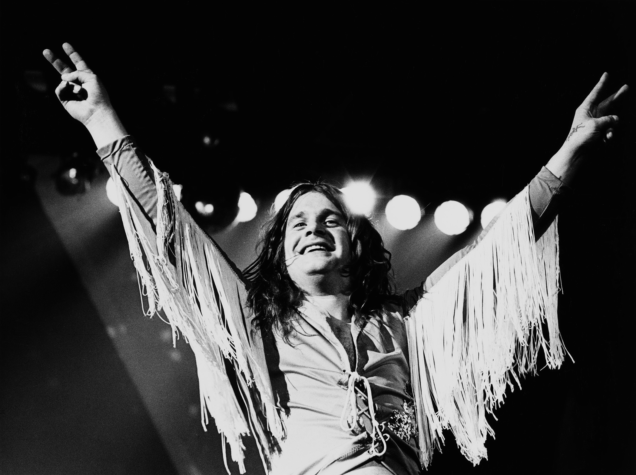 Ozzy Osbourne throws a double peace sign live onstage wearing his white tasseled jacket.