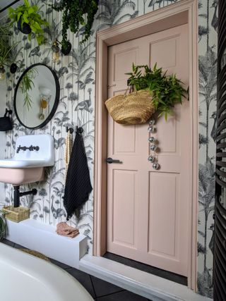 Baby pink bathroom door with feature nature-inspired wallpaper, hanging plants and round mirror