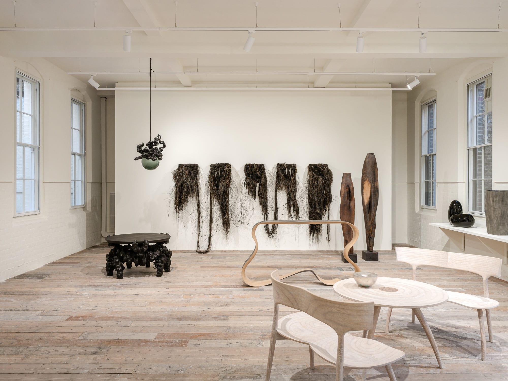 A white-painted gallery with pale wooden floors holds a series of fiber-based installations hanging from its walls, alongside wooden sculptures and furniture.