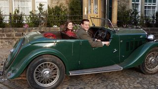 Rachel Shenton as Helen in a red coat and Nicholas Ralph as James in a tweed suit sit in a green car outside the veterinary practice.