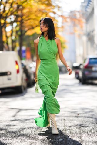 guest wears gold earrings, a green halter neck / sleeveless / wrap long dress, a beige fabric Chiquito handbag from Jacquemus, pale yellow shiny leather heels knees boots / high boots , outside Awake, during Paris Fashion Week - Womenswear Spring/Summer 2023, on October 04, 2022 in Paris, France