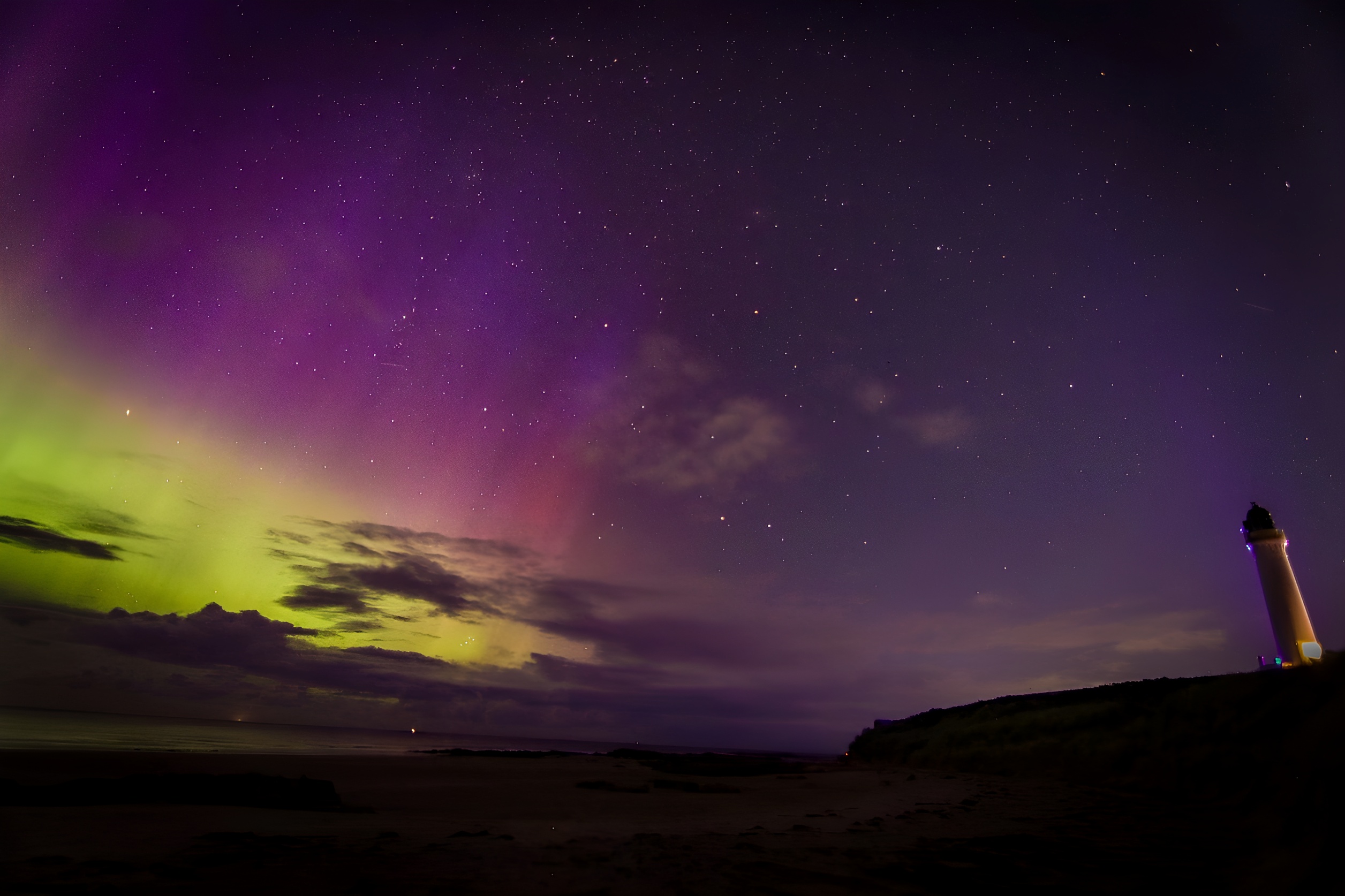 Zielono-fioletowa zorza polarna na niebie z małą latarnią morską po prawej stronie zdjęcia.