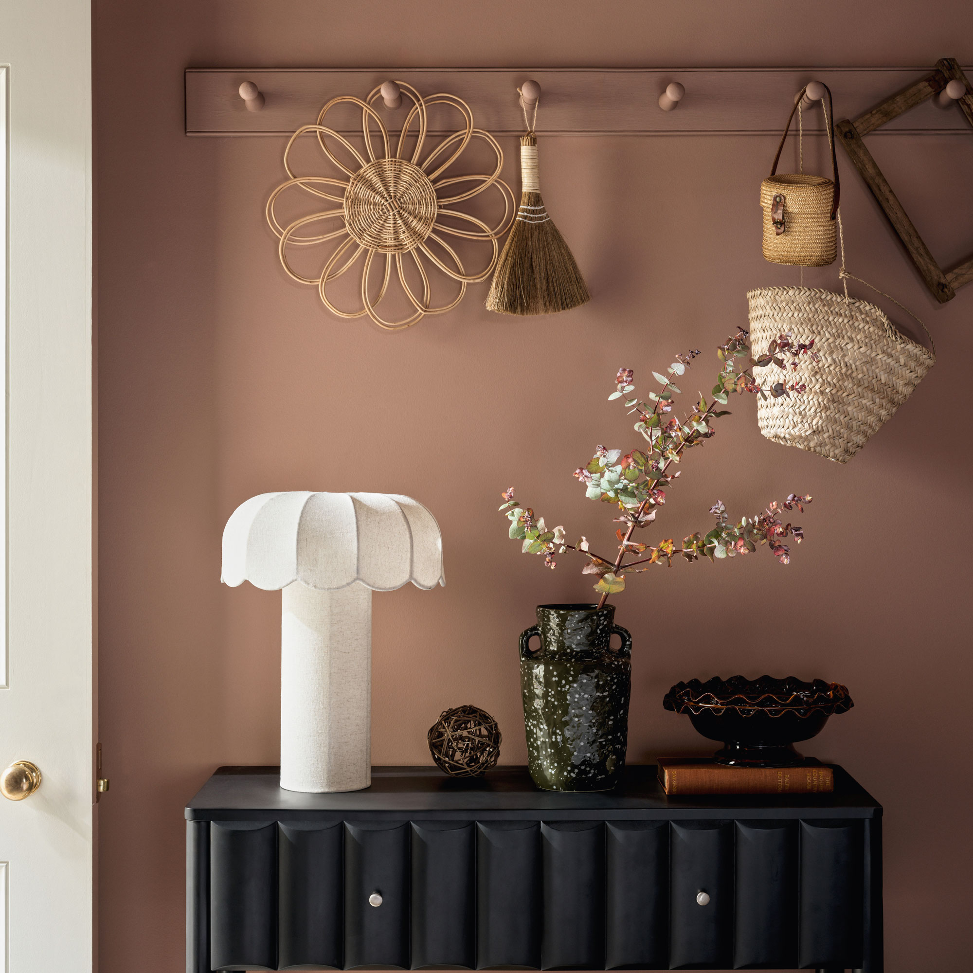 Dusty pink background with black console table, white skeletal table lamp, wooden hooks and straw accessories.