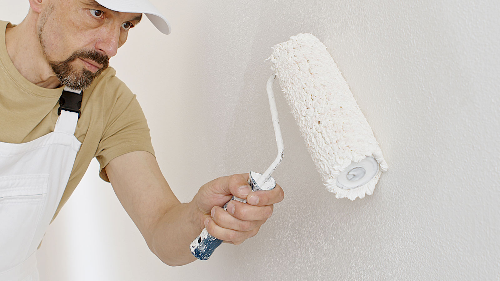 Man in white overalls, cap and light brown tee shirt rollering a wall with white paint