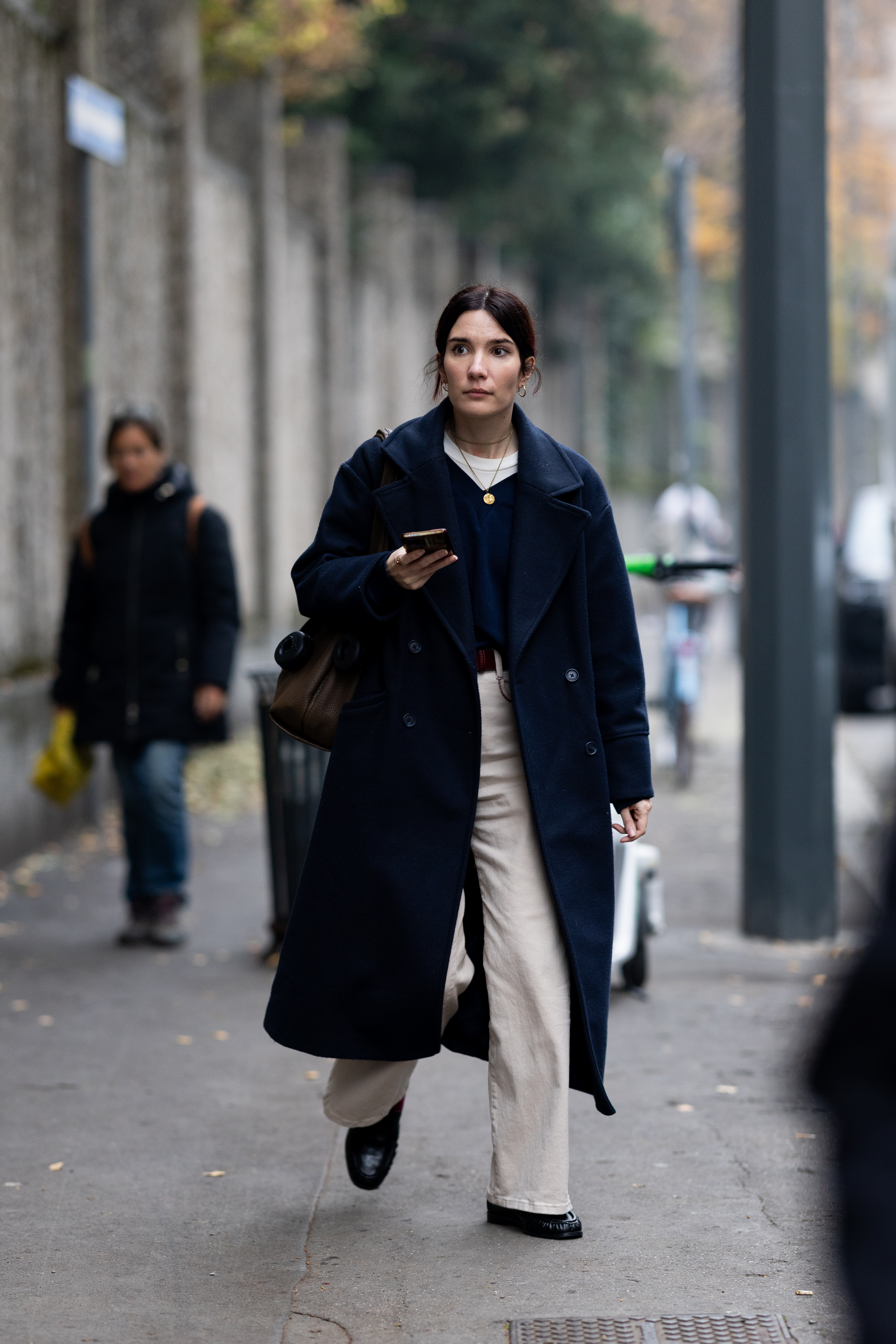 A passerby wears a long navy double-breasted coat over a dark sweater and beige wide trousers, paired with black loafers, gold jewelry, and a brown structured bag outside Grazialand event on November 14, 2025 in Milan, Italy. (Photo by Valentina Frugiuele/Getty Images)