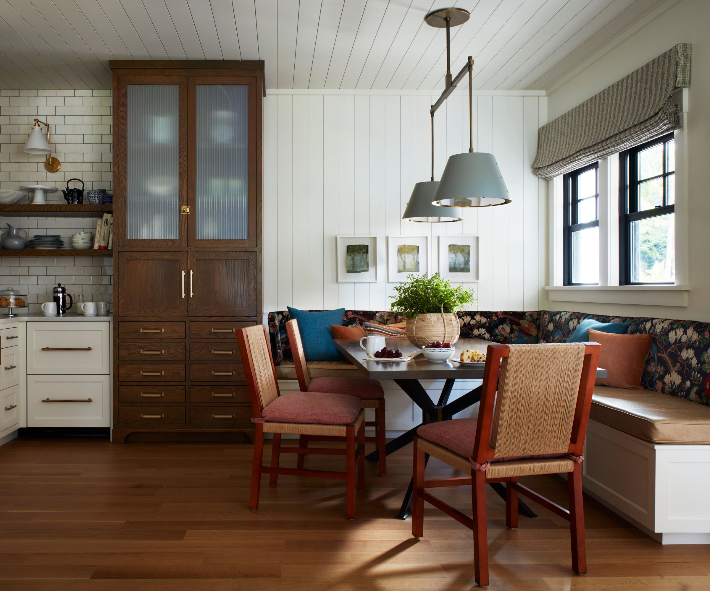 white farmhouse kitchen with panelled walls, a large dark oak pantry cupboard and a large banquette breakfast nook with floral upholstery, leather and wooden chairs and blue scatter cushions and two light blue pendant lights