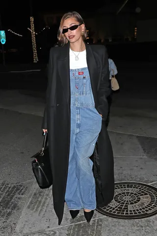 Hailey Bieber wearing long overalls, black slingback heels, and black long coat.