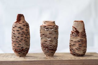 3 Sculptured vessels in shades of brown with unevenly placed oval shaped holes on the body, placed on a wooden table and photographed againt a white background